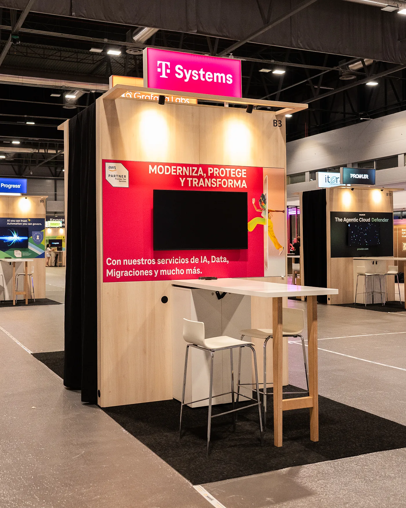 T-Systems stand on the exhibition floor, with a magenta print wall, a screen and a high table with stools