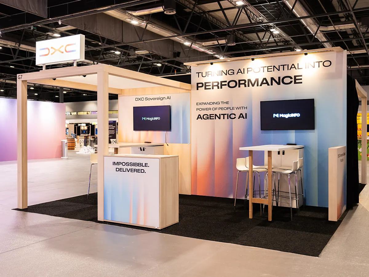 DXC stand with a light print wall in blue and orange, a screen and a high table with stools