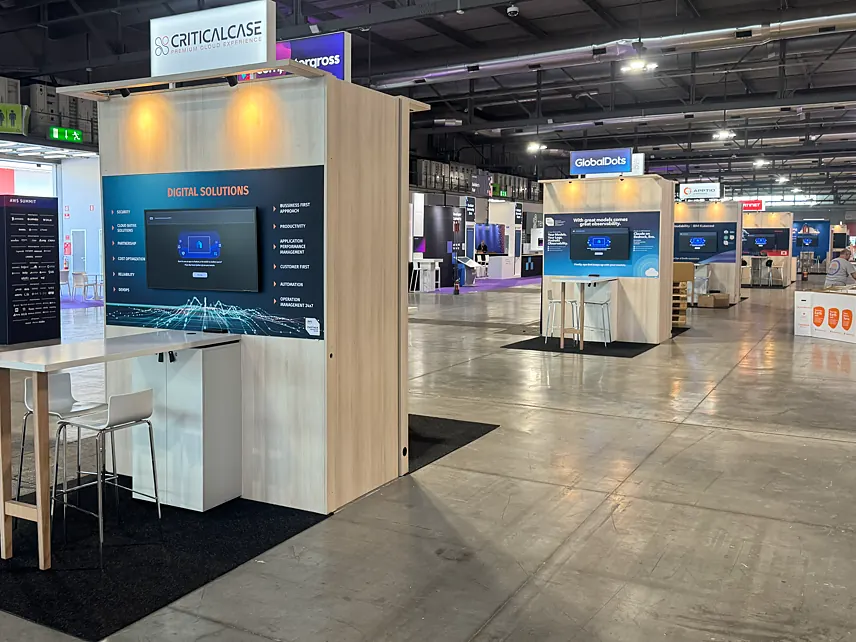 Criticalcase stand with a dark blue print wall, a screen and a high table with stools on the exhibition floor